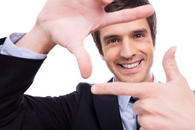 Focus on me! Playful young man in formalwear gesturing finger frame and looking through it while standing isolated on white background 